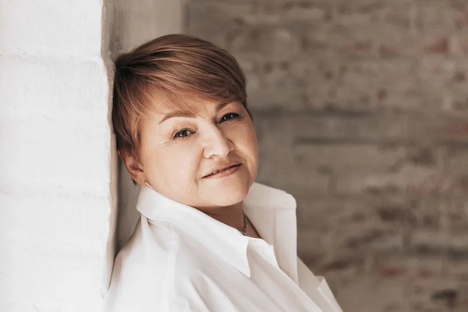 Charming senior woman poses in a stylish white shirt against a textured wall, exuding confidence and elegance.