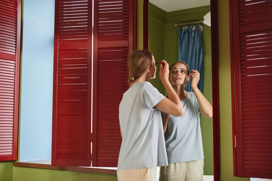 Woman applying mascara in front of a mirror with red shutters reflecting her image indoors.