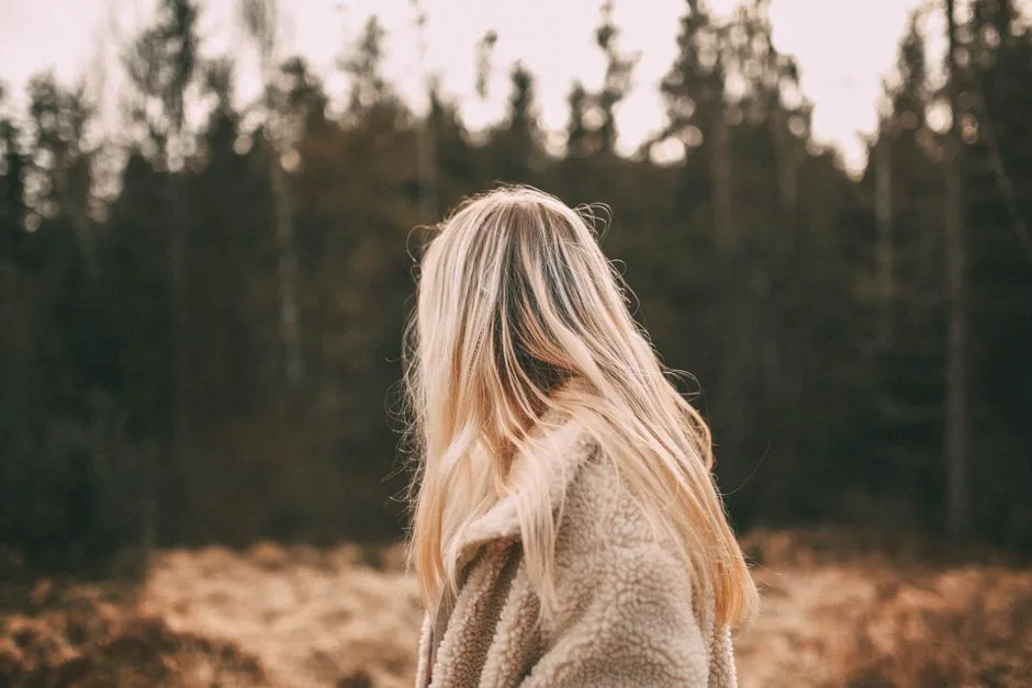 Blonde woman in a coat standing in a forest during autumn, captured from behind.