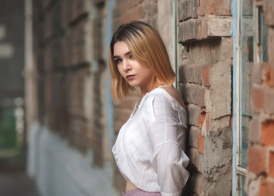 Model with blonde hair in white top leaning against a brick wall, showcasing modern fashion.