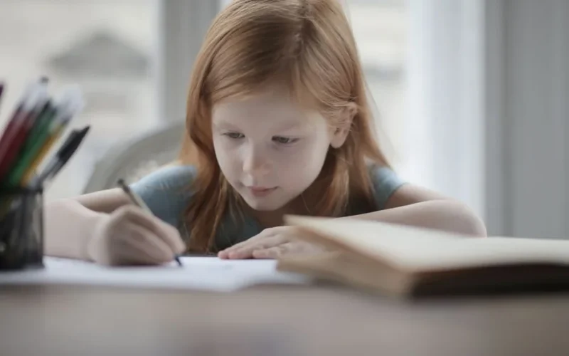 Adorable positive preschool child with red hair in casual wear sitting at table with book and pens or pencils while making exercise on paper sheet against blurred window