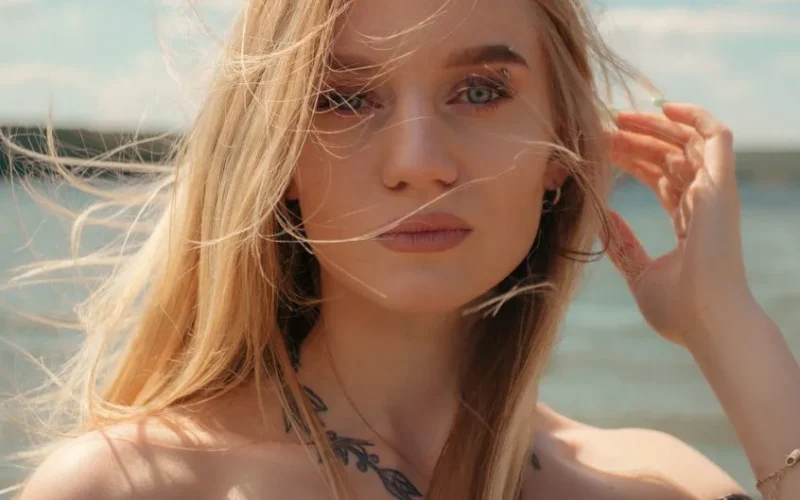Blonde woman with tattoos enjoying a breezy day by the lake, captured in a fashion portrait.