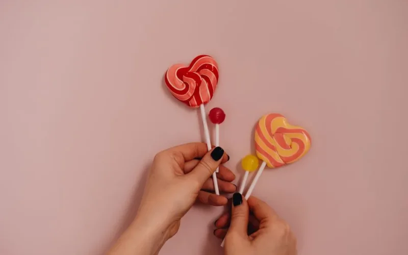 Close-up of hands holding colorful heart-shaped lollipops on a pink background.