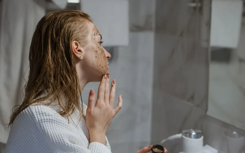 Woman with wet hair using coffee scrub on face in a modern bathroom setting.