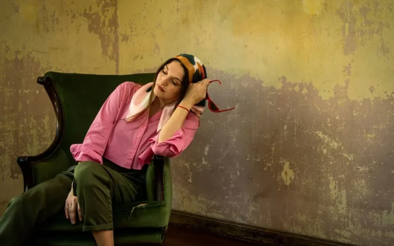 Elegant woman with silk scarf seated on a vintage green chair in a rustic room.