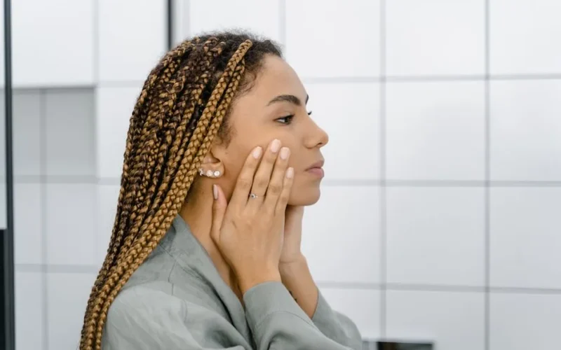 Profile view of a woman with braided hair gently touching her face, standing in front of a mirror.