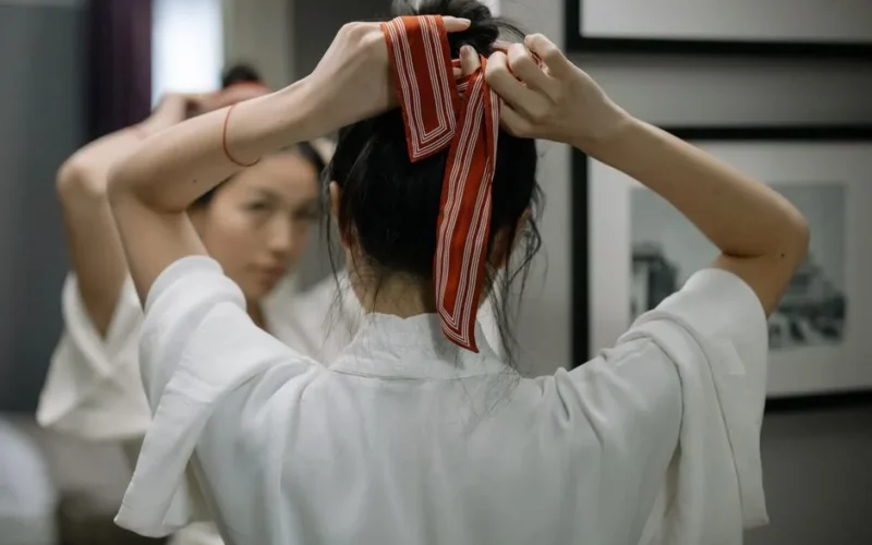 Close-up of a woman in a robe tying her hair with a red ribbon in front of a mirror indoors.