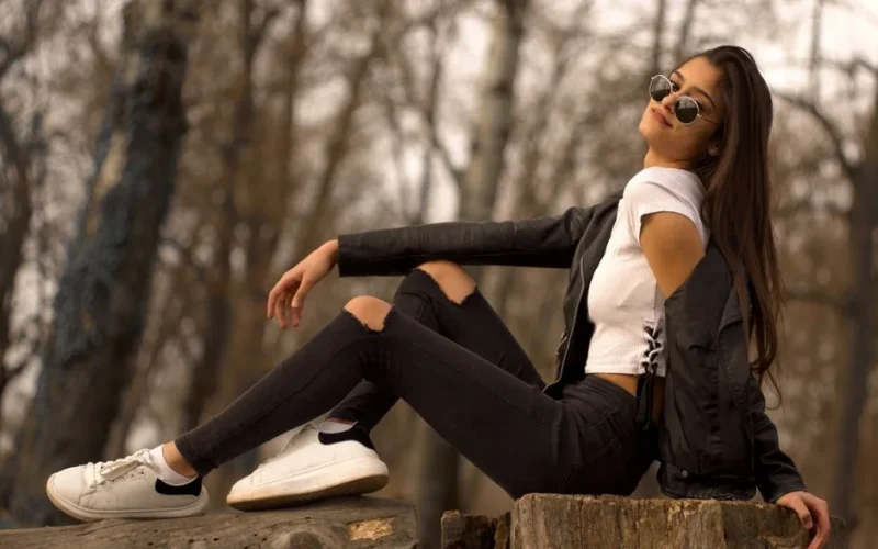 Stylish young woman in sunglasses and leather jacket posing outdoors on a tree stump.