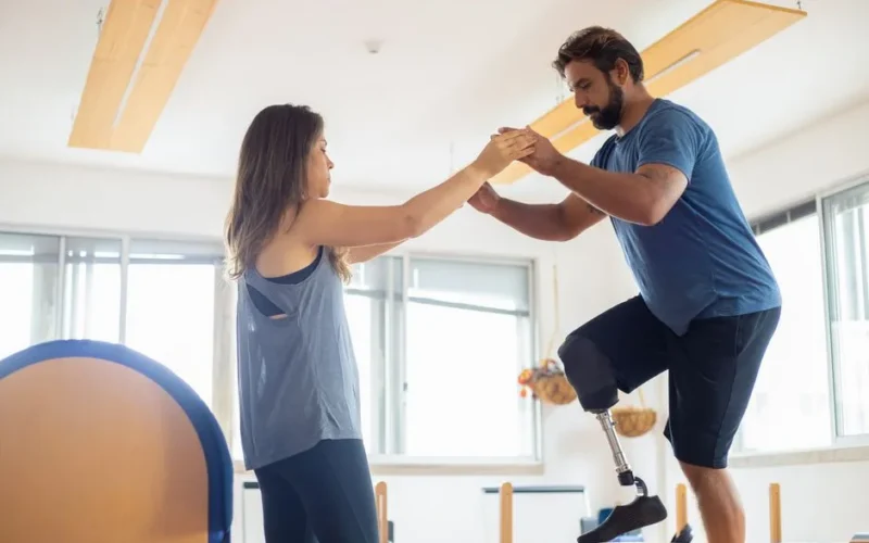A man with a prosthetic leg exercises with a therapist in a well-lit gym environment.