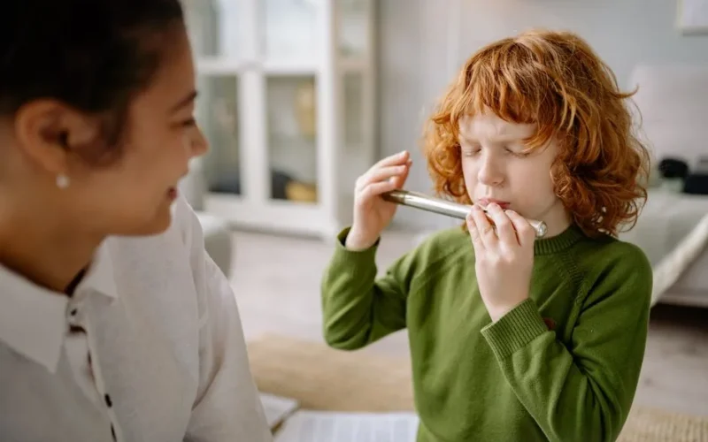 A young boy with red hair plays a flute indoors while his mother smiles nearby.