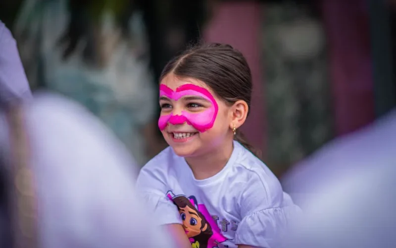 A young girl with vibrant pink face paint smiles joyfully outdoors.