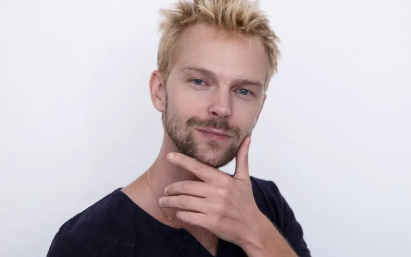 Portrait of a confident blonde man posing with hand on chin against a white background.