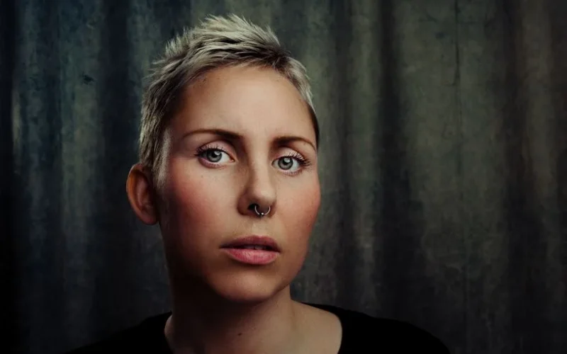 A striking portrait of a woman with short blonde hair, a nose piercing, and a contemplative expression.