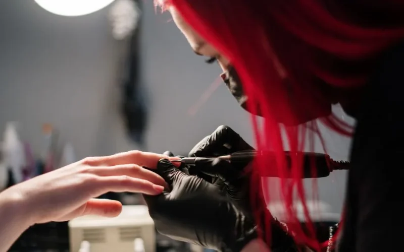 A nail technician with red hair performs a manicure wearing black gloves in a salon.