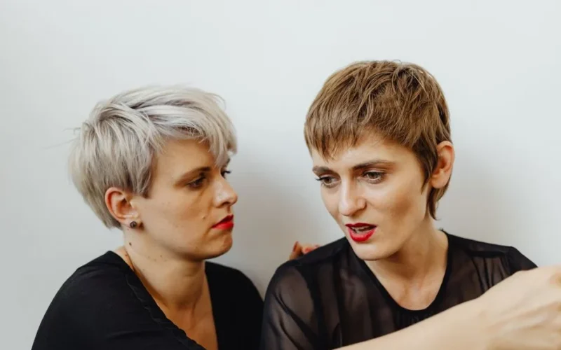 Two women with short hairstyles having an intense discussion indoors against a white wall.