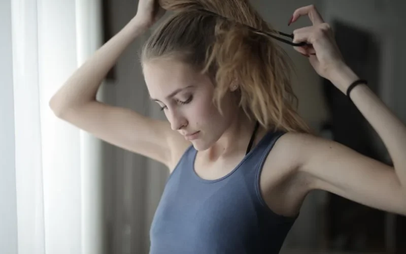 Young tranquil female in casual clothing standing near window with curtain in living room and tying ponytail while resting at home during weekend
