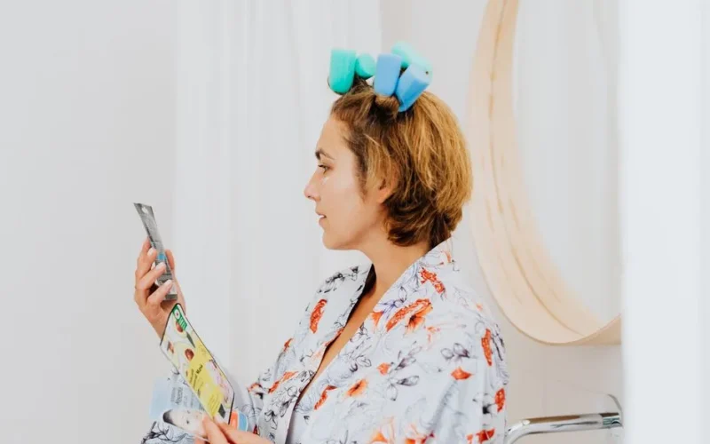 Woman in a bathrobe with hair curlers reads skincare instructions indoors.