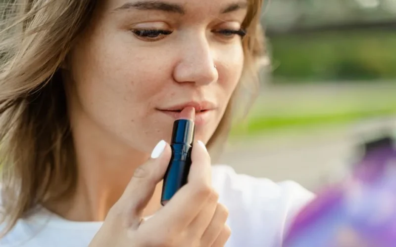 Close-up of a woman applying lipstick outdoors with a natural background.