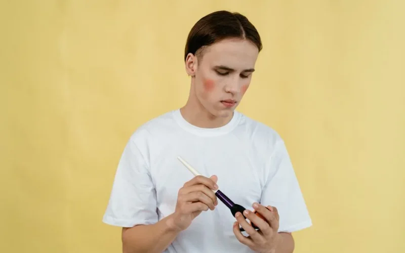 Portrait of a young man applying blush using a makeup brush with a yellow backdrop, wearing a white shirt.