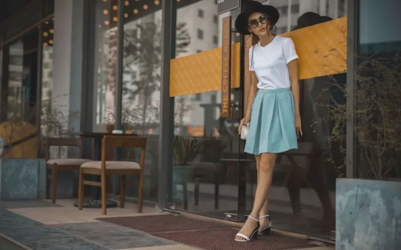Stylish woman wearing a white shirt and blue skirt, accessorized with a hat and sunglasses, standing outdoors.