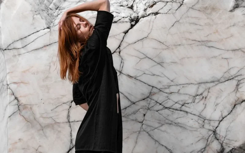 Elegant redhead woman posing in stylish black outfit against a marble wall.