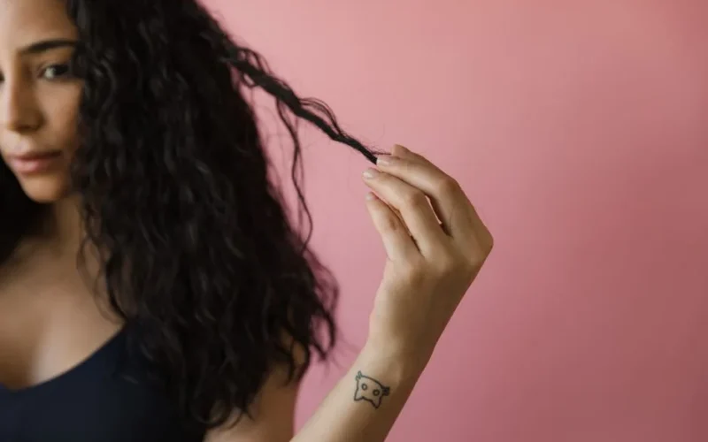 A woman with tattooed wrist examines her curly hair against a pink background.
