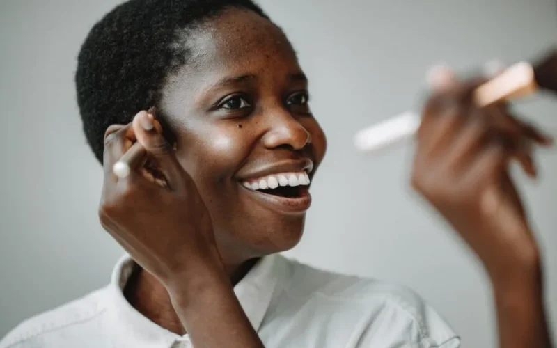 A joyful woman applying makeup with a brush in front of a mirror indoors.