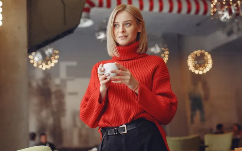 Blonde woman in cozy café wearing red sweater, holding a ceramic cup.