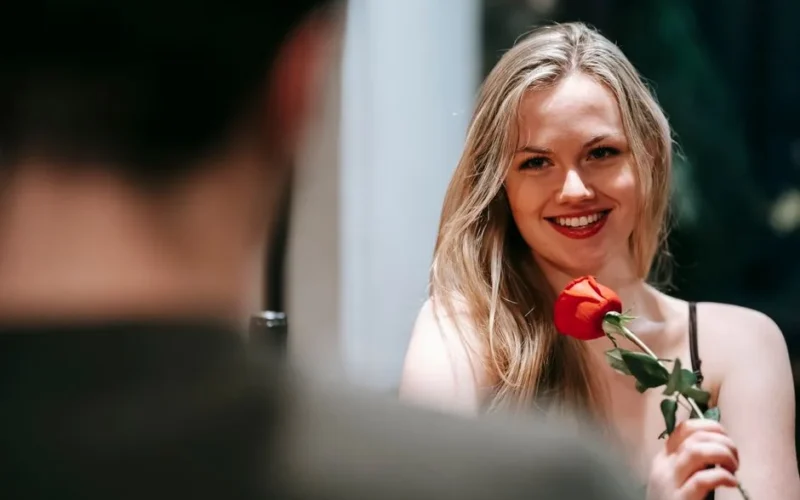 Young couple in elegant clothes sitting in light restaurant while enjoying date with red rose and looking at each other