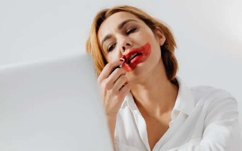 Young woman playfully applying red lipstick, creating an artistic makeup effect.