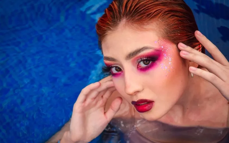 Captivating portrait of a woman in water, featuring striking red hair and bold makeup against a blue background.