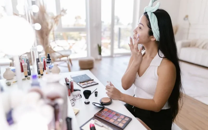 A woman applies makeup at a vanity in a well-lit room, surrounded by cosmetics.