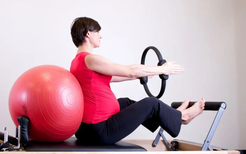 A pregnant woman in a red top practicing Pilates using a fitness ring and exercise ball in a gym setting.