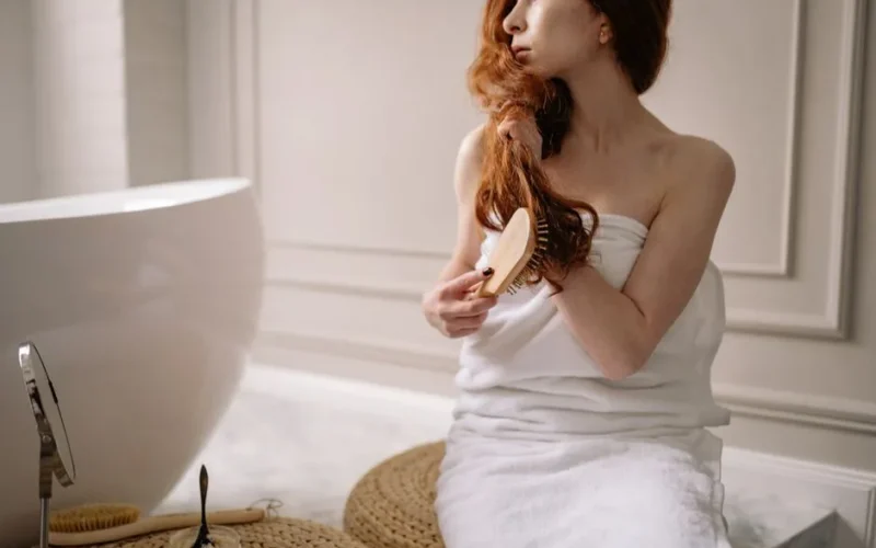 Woman wrapped in towel brushing hair in a modern indoor bathroom setting.