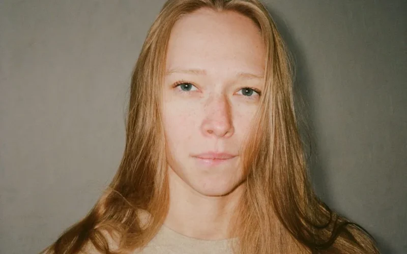 A serene close-up portrait of a woman with blond hair and freckles indoors.