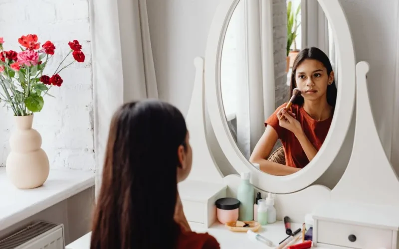 A teenage girl applies makeup using a brush while looking in a mirror indoors.