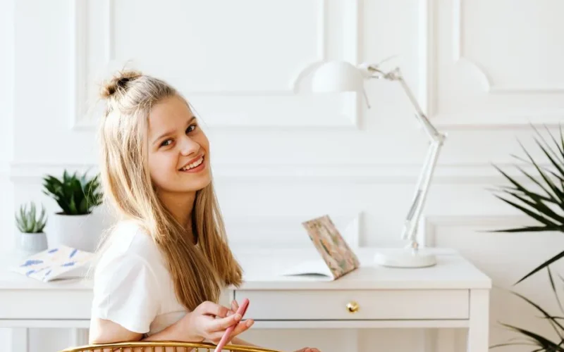 Cheerful teenage girl holding a pencil while studying at a desk in a bright, modern room.