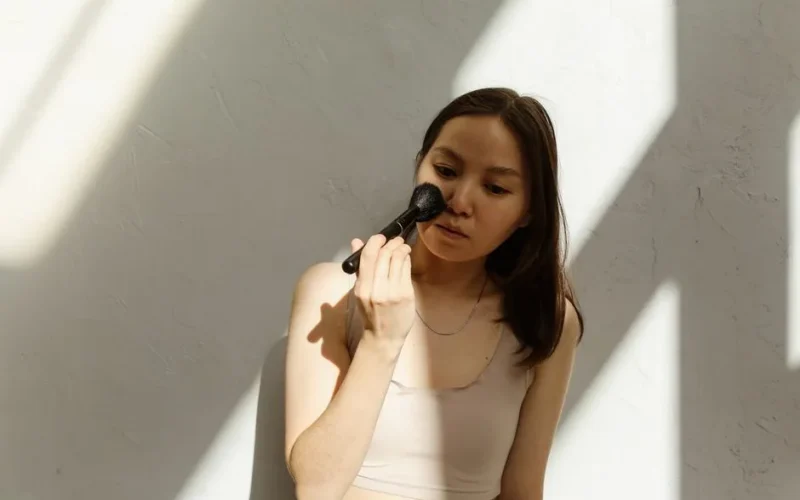 Portrait of an Asian woman applying makeup with a brush, bathed in sunlight.