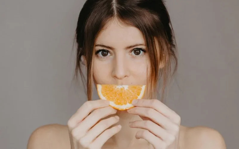 Close-up portrait of a woman holding a half orange slice, with a focus on natural beauty and skincare.