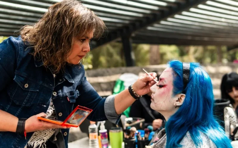 A makeup artist expertly applies fantasy makeup to a woman with vibrant blue hair outdoors.