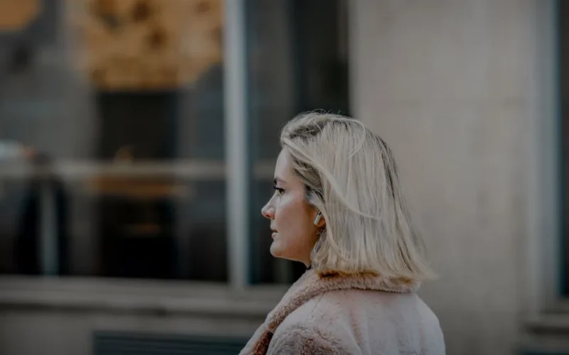 Profile of a stylish blonde woman walking outdoors in an urban environment, wearing a coat.