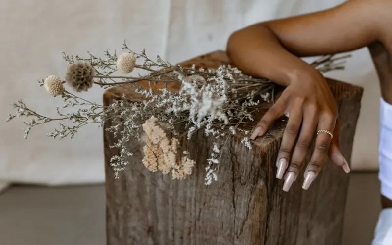 A delicate hand with manicured nails and silver ring rests on a rustic wooden block adorned with dried flowers.