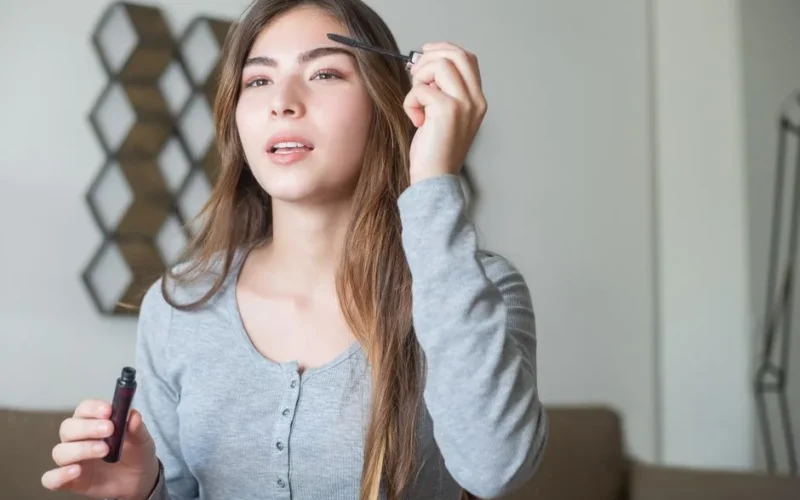 A young woman applying eyebrow makeup with a brush indoors.
