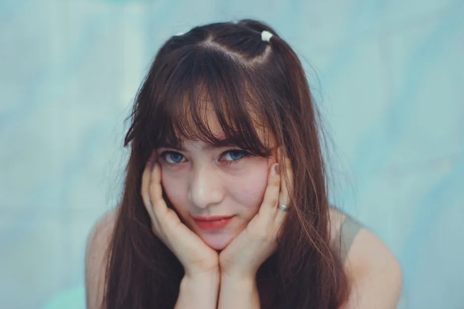 A close-up portrait of a young woman with hands on cheeks and a gentle expression.