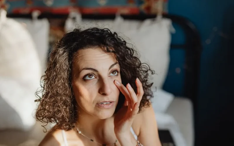 Woman applying skincare in a sunlit bedroom, embracing morning relaxation and self-care.