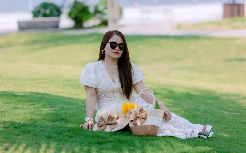 Woman in a white dress sitting on grass with woven bags, enjoying a sunny day.