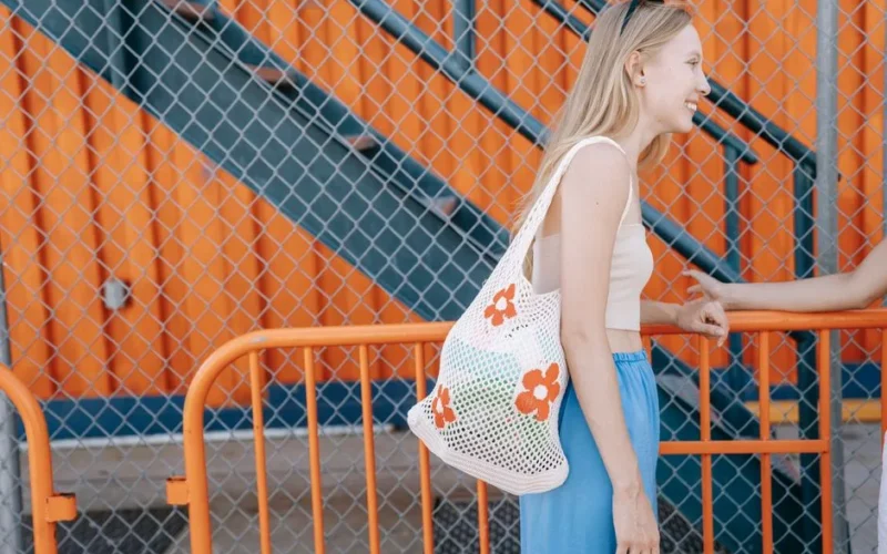 Casual outdoor portrait of a smiling woman leaning on a metal railing with a floral tote bag against a vibrant orange background.