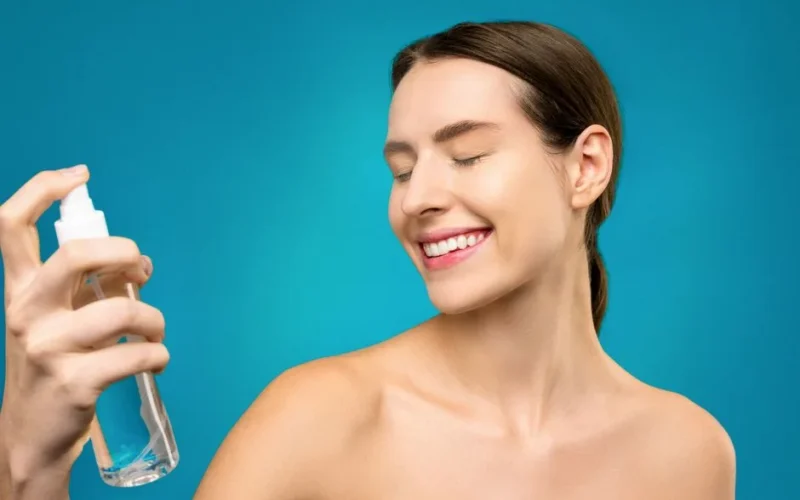 Smiling woman applying a refreshing facial spray for skincare on a vibrant blue background.