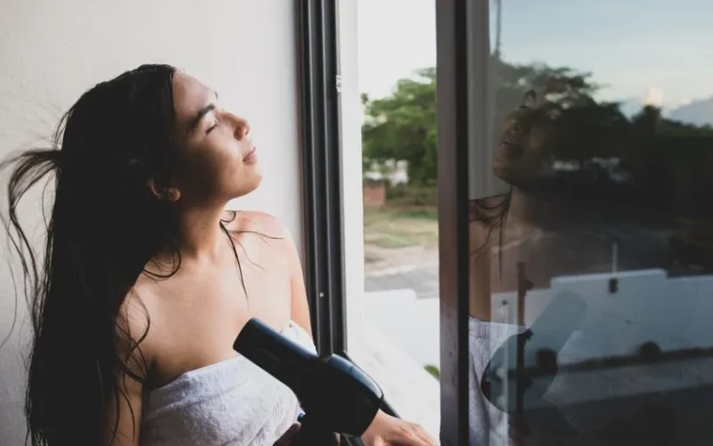 Young woman drying hair with blow dryer by open window at home.