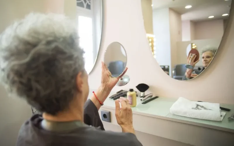 A senior woman inspects her new hairstyle at a salon in Portugal, reflecting in a handheld mirror.